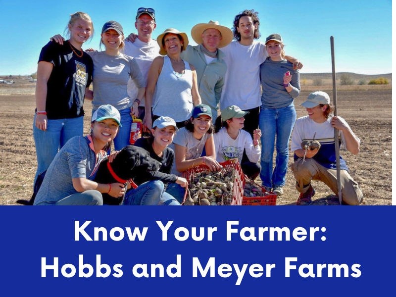 A group of ten smiling people, likely farmers, pose with a dog and harvested produce in a field under a sunny sky.