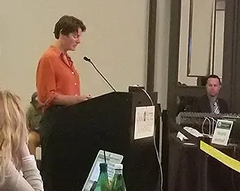 Marian Blom with short brown hair wearing an orange shirt, is speaking at a podium with a microphone.