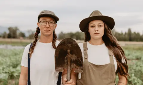 Two female farmers standing side by side in a field, with the woman on the left wearing glasses and holding a shovel, while both wear hats.
