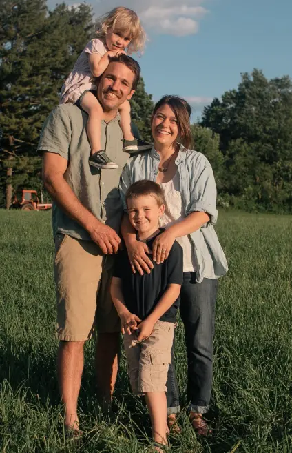 A happy family of four posing together in a lush green field on a sunny day, with a tractor visible in the background and trees lining the horizon under a clear blue sky.