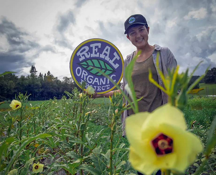 A smiling woman wearing a black cap and gray shirt stands in a field of okra plants with yellow flowers, holding a circular sign that reads 'Real Organic Project' with a green leaf logo.