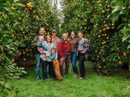 A multi-generational family of farmers posing together in an orange grove, surrounded by lush green trees laden with ripe oranges, representing a family-owned organic farm.