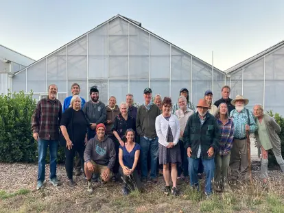 A diverse group of approximately 19 people posing together in front of two large greenhouses, with some kneeling in the front row and others standing behind them, dressed in casual clothing suitable for farming or gardening.