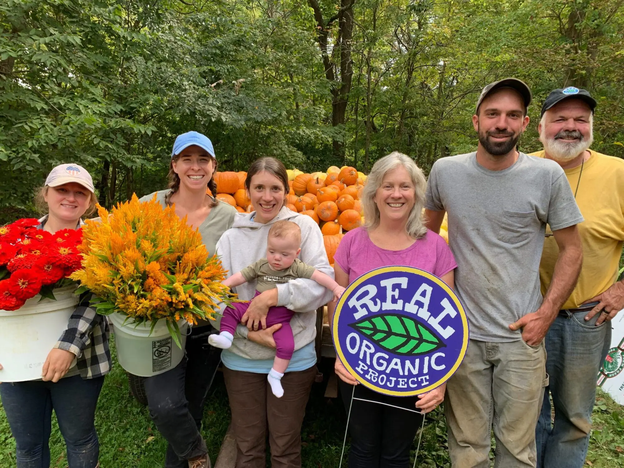 Farm team and supporters holding organic flowers and a Real Organic Project sign in front of harvested pumpkins.