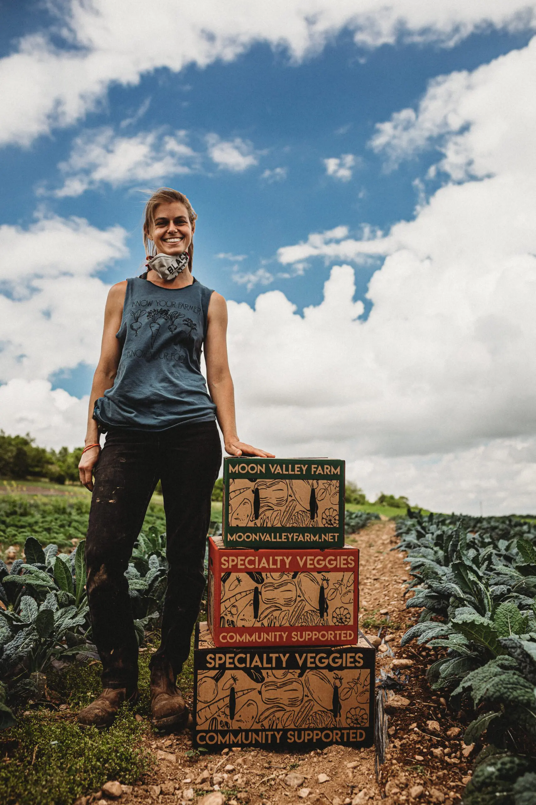 Farmer from Moon Valley Farm standing in an organic kale field with boxes of community-supported specialty veggies, promoting local farms.