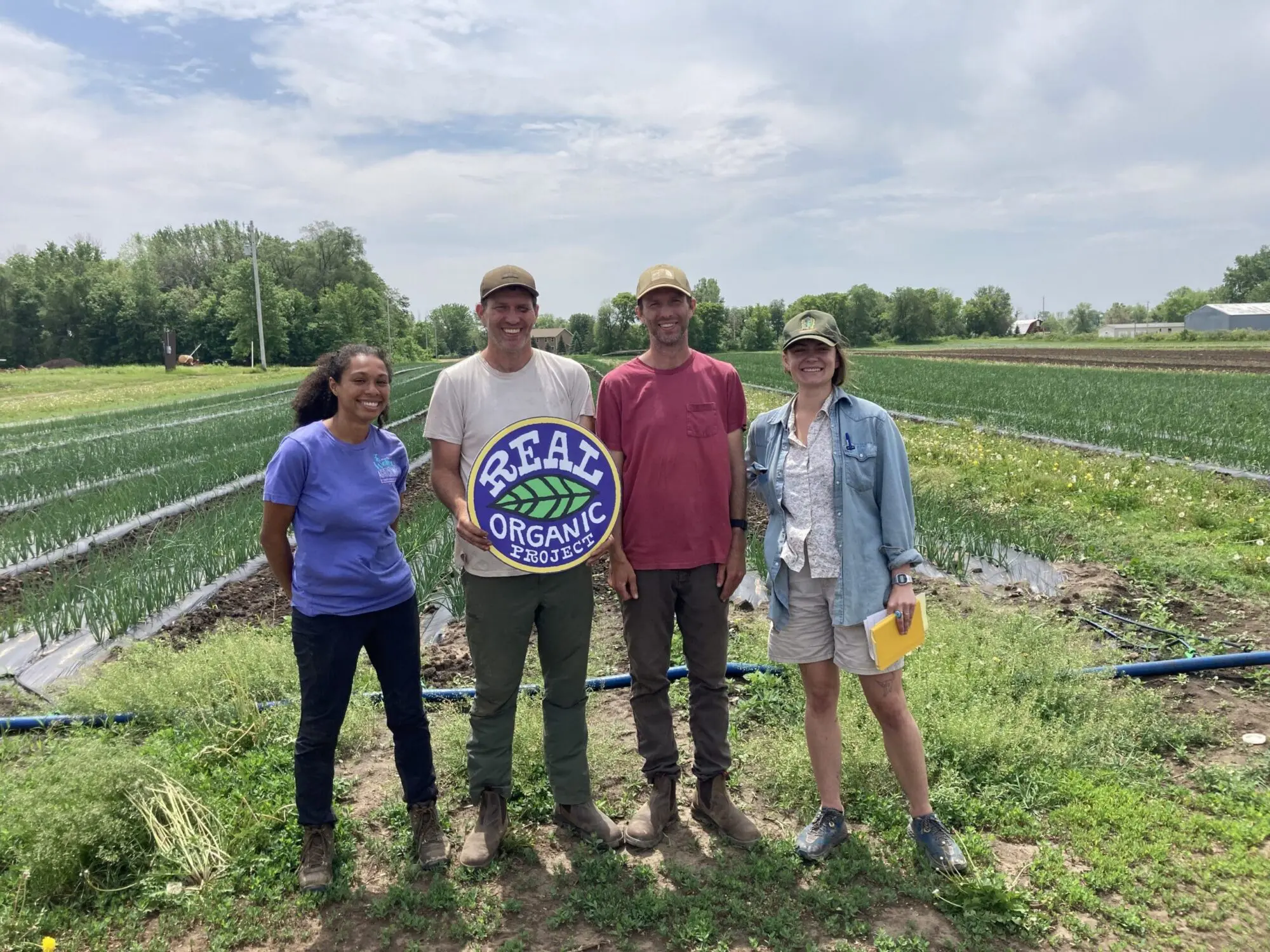 Farmers holding a Real Organic Project sign in an organic field with rows of onions, practicing regenerative farming.