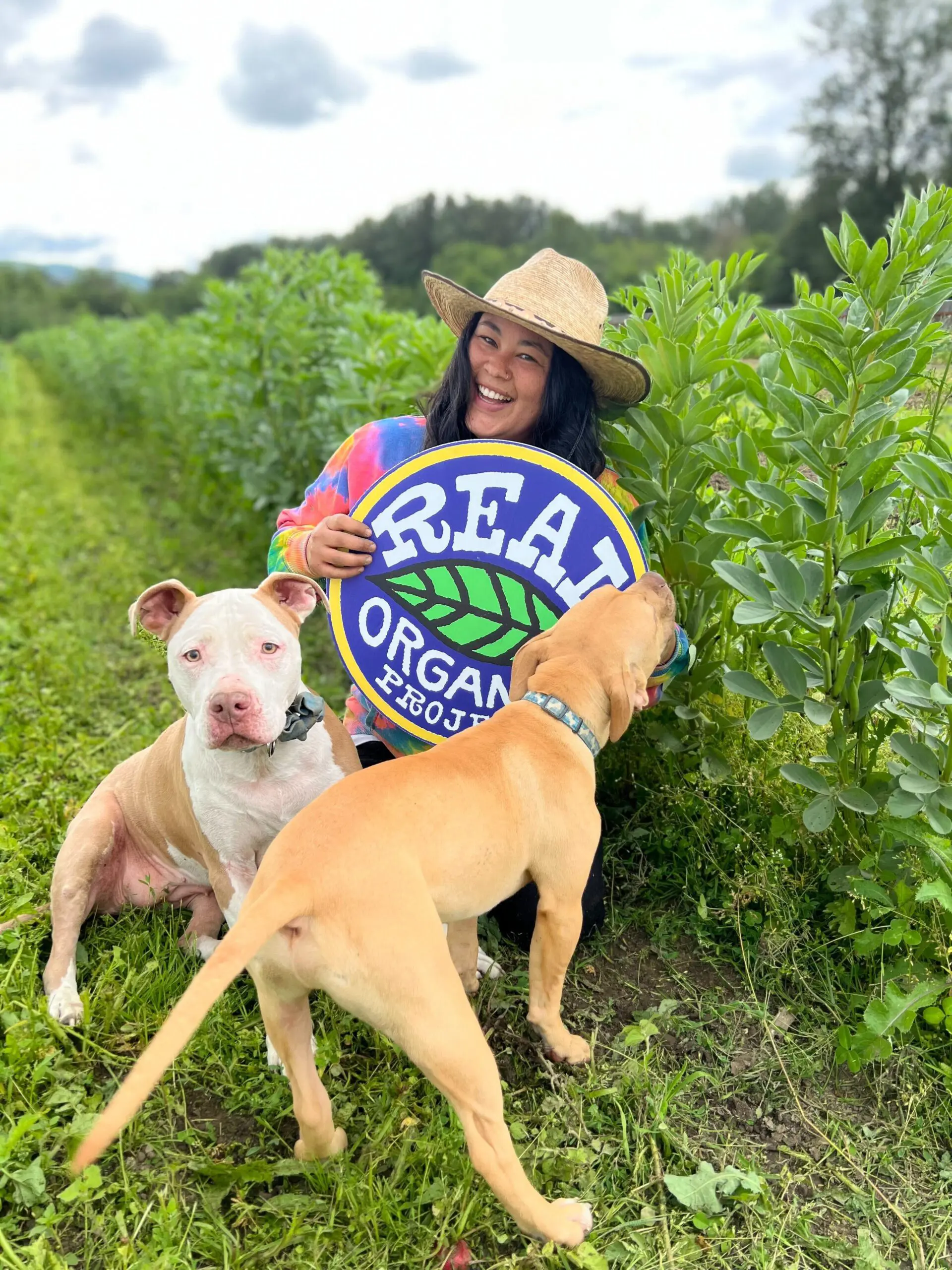 Farmer holding a Real Organic Project sign surrounded by dogs in an organic field.