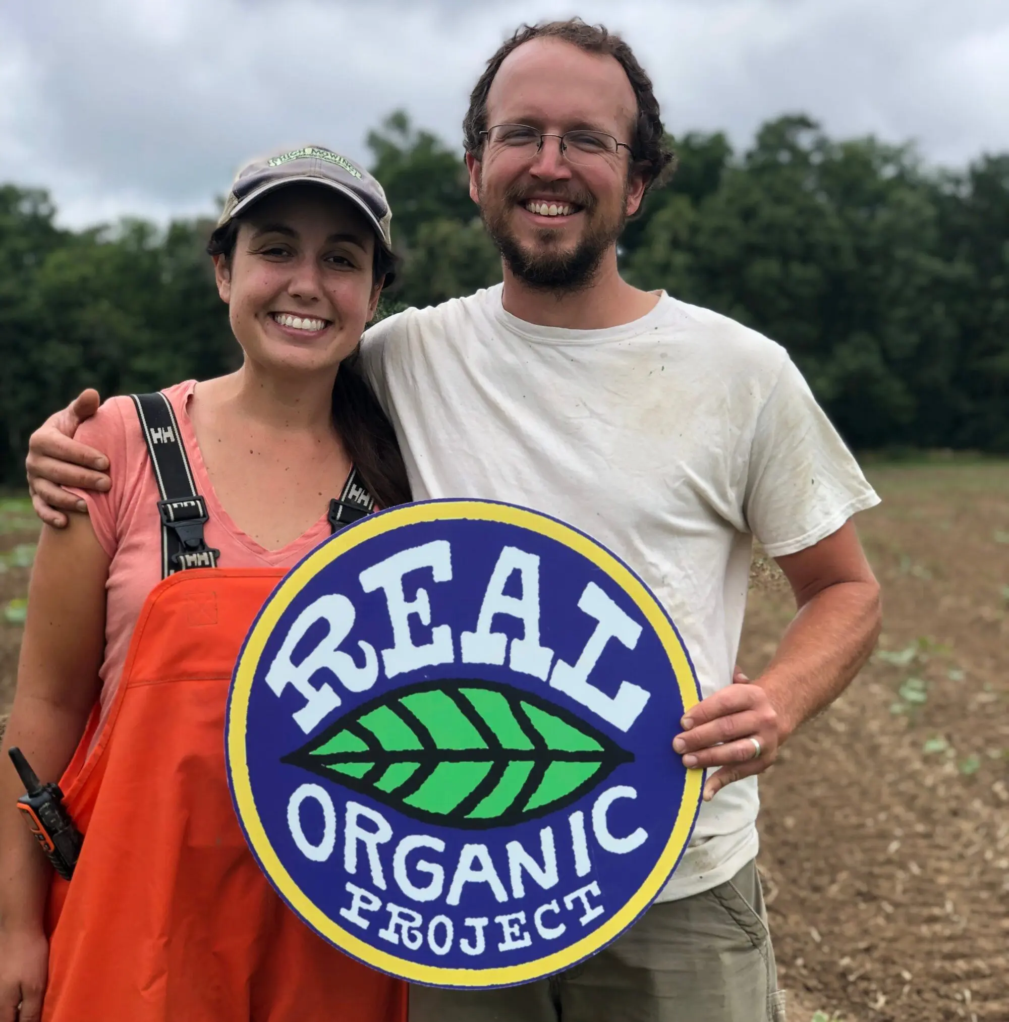 Smiling farmers holding a "Real Organic Project" sign in a field.
