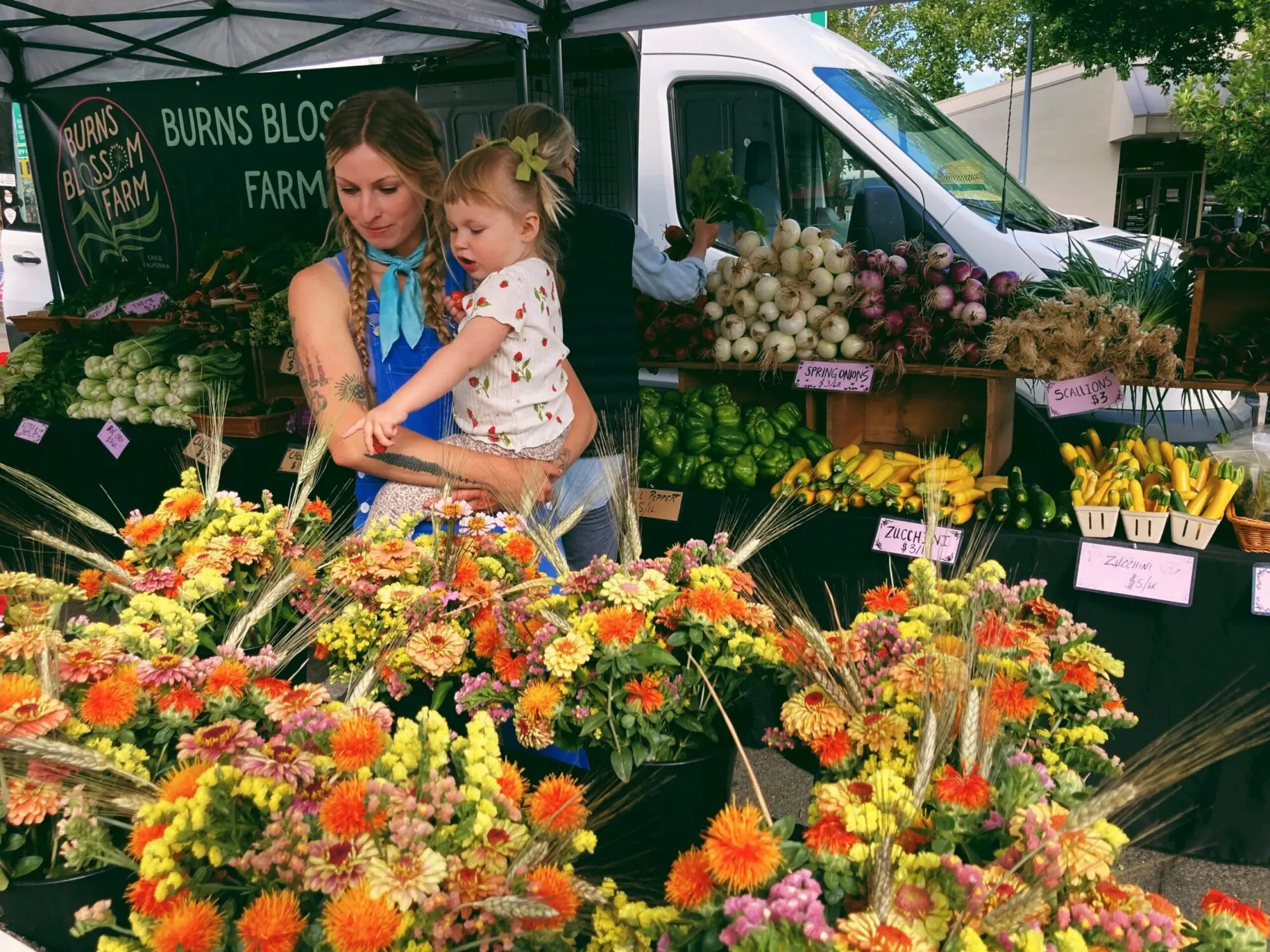 Farmer and child at a vibrant farmers market stand with organic vegetables and flowers.