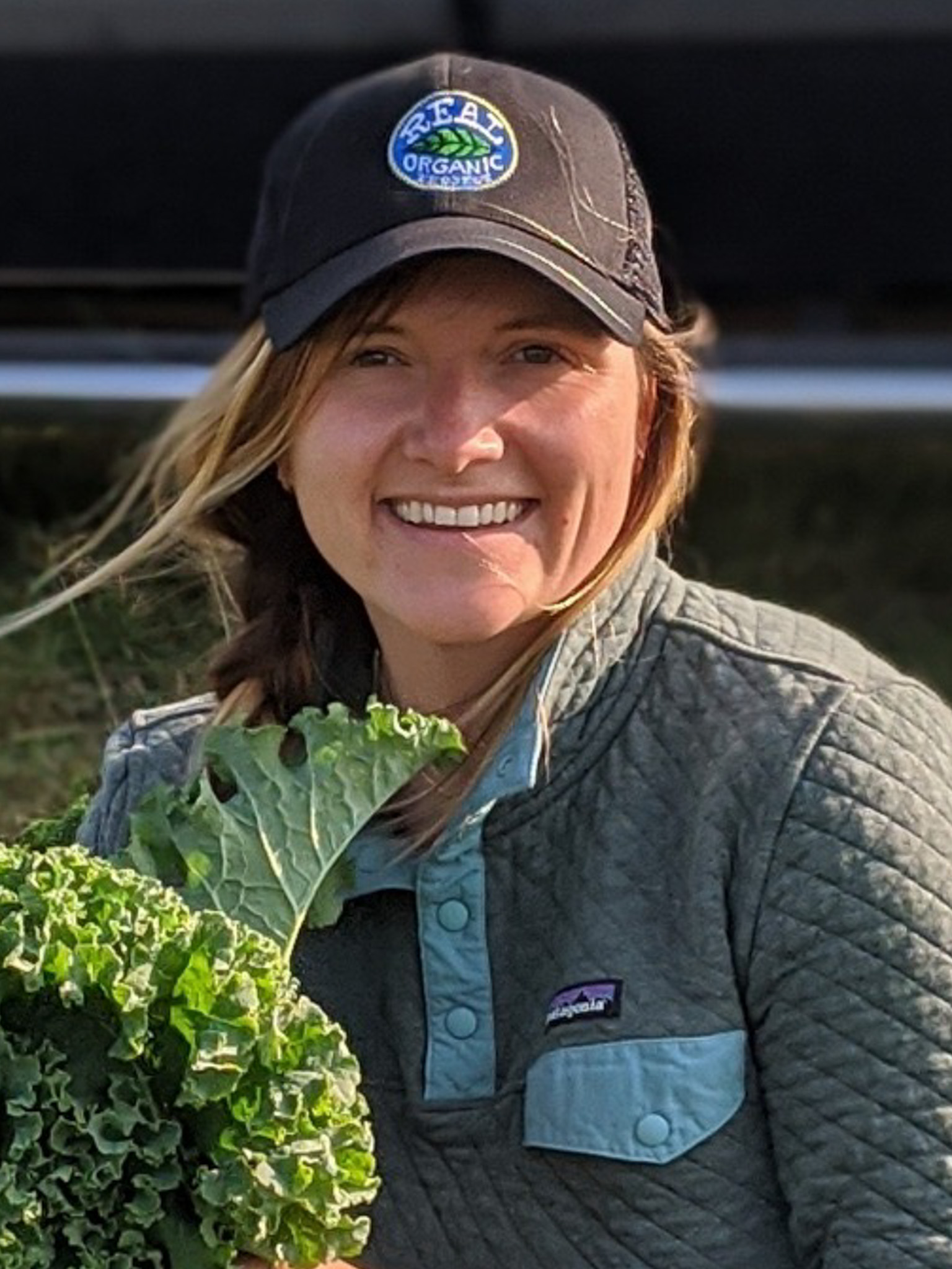 Anaïs Beddard, white lady with blonde hair, smiles with a leaf of curly kale and real organic project hat on.