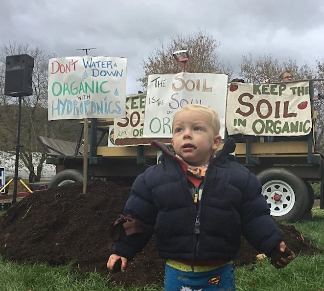 A child stands in front of a pile of soil and tractors at a "Keep Soil In Organic" Rally.