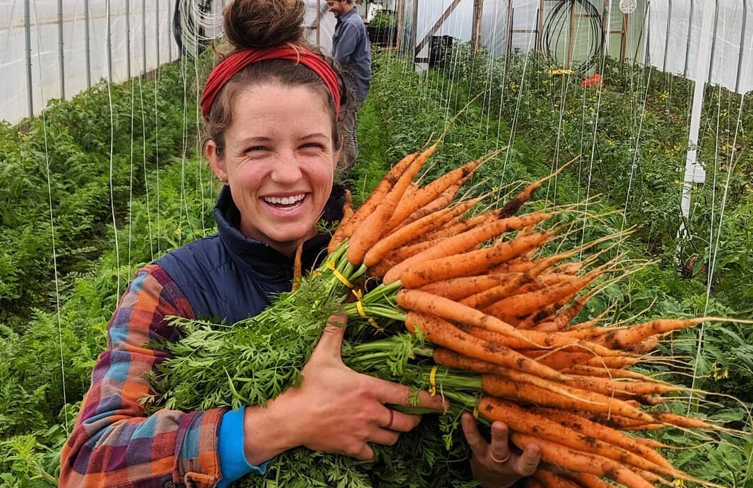 Footprint Farm farmer holding a large bundle of carrots and smiling in a high tunnel.