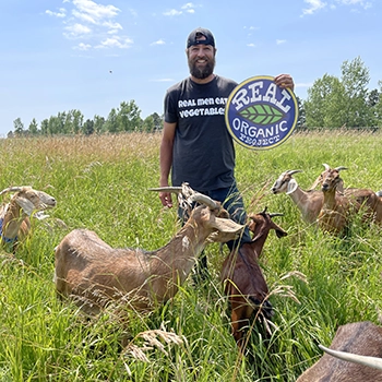 farmer in pasture surrounded by goats holding a Real Organic Project sign and wearing a t shirt that says real men eat vegetables