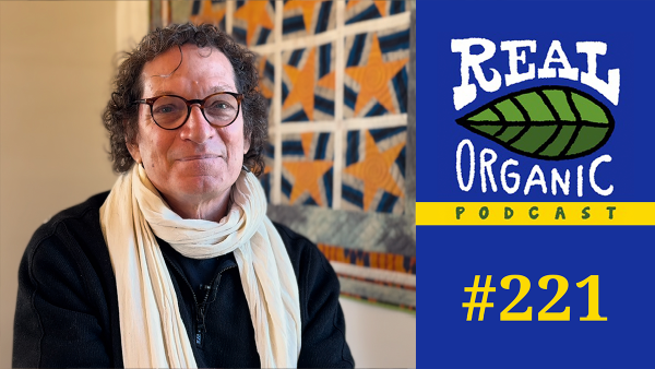 Organic Rising filmmaker Anthony Suau seated with black-framed glasses, a black shirt and a cream-colored scarf around his neck.