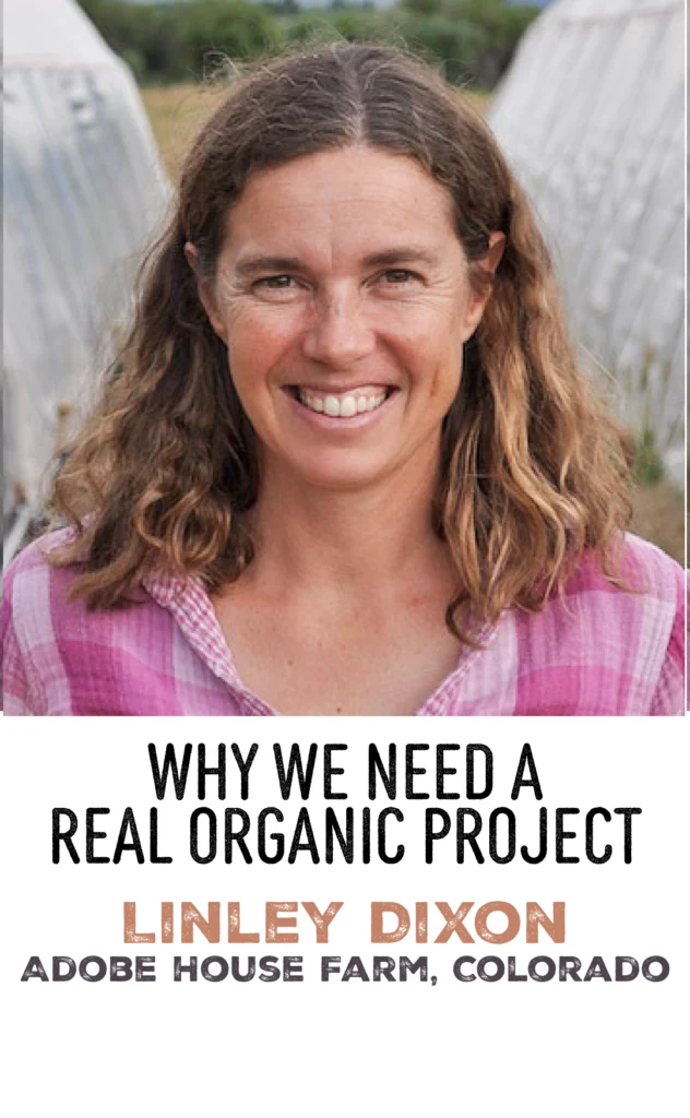 Real organic project farmer Linley Dixon smiles in a pink plaid shirt near a greenhouse